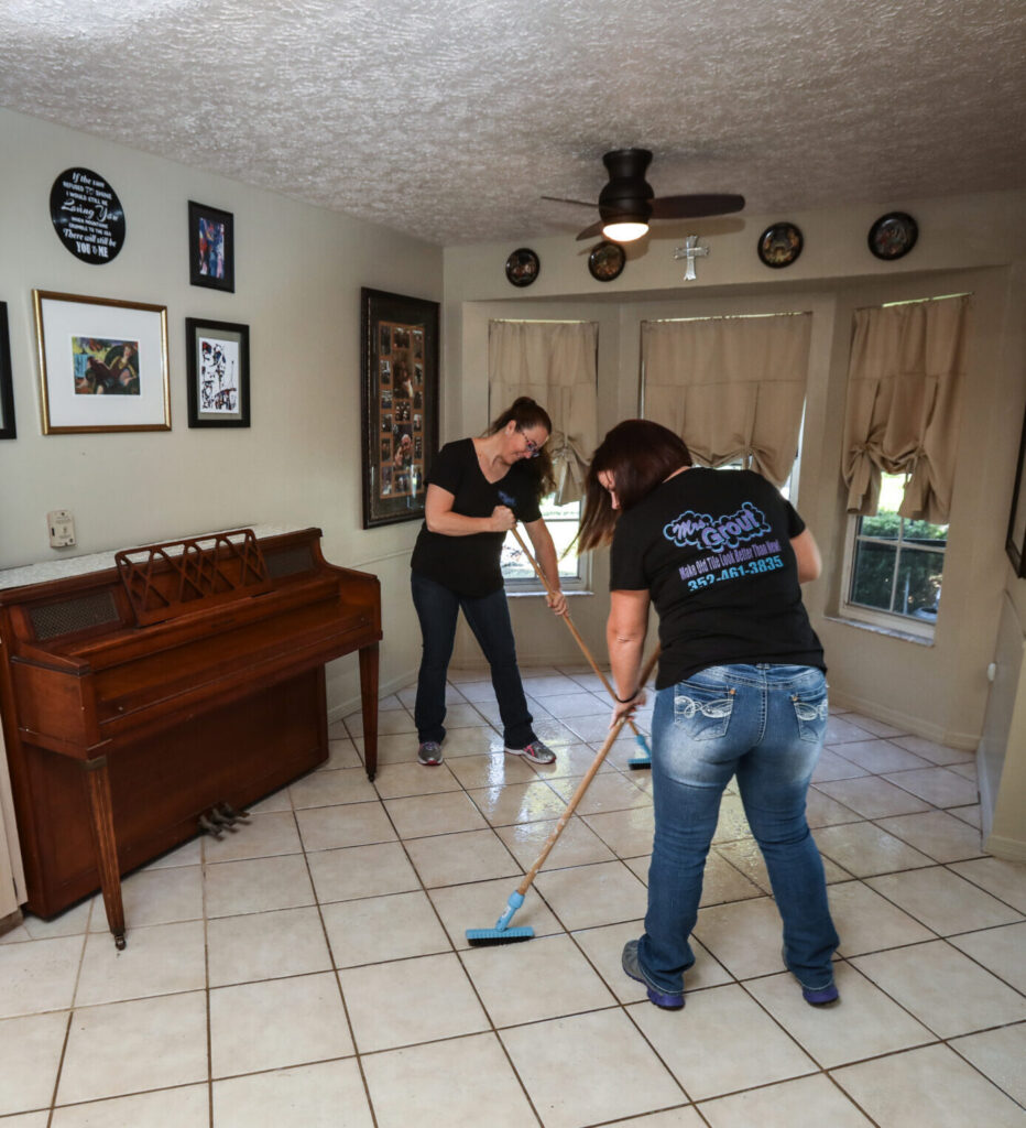 Two Mrs. Grout team members clean and restore grout lines on a tiled floor inside a home using professional cleaning tools. The image highlights the team's tile and grout restoration process in a residential setting.