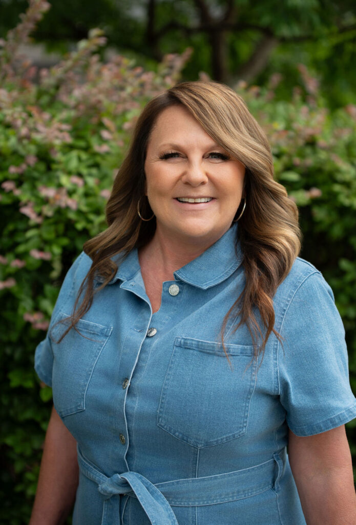 Portrait of Lisa Council, owner and operator of Mrs. Grout, smiling outdoors in front of green landscaping. The image introduces the owner and leader of the company.