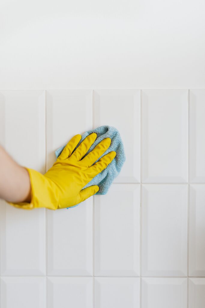 Close-up of a person wearing a yellow cleaning glove wiping white wall tiles with a blue microfiber cloth. The image shows routine tile cleaning and surface maintenance.