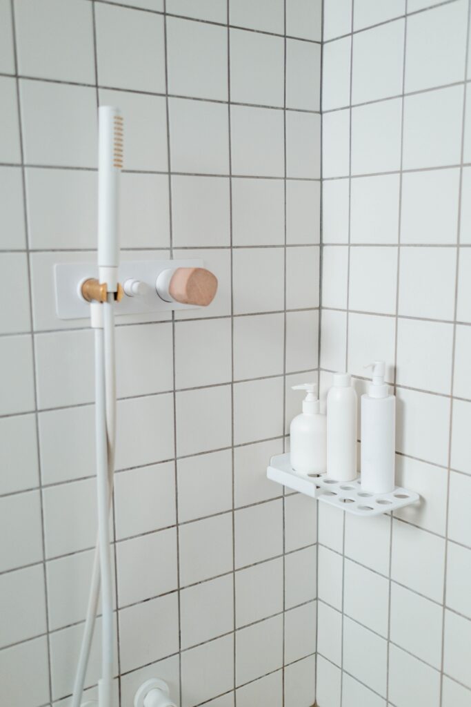 Clean white tiled shower with dark grout lines, a handheld showerhead, and a wall-mounted shelf holding three soap dispensers. The image highlights a clean, organized shower space.
