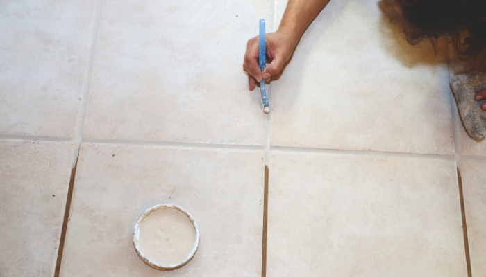 Mrs. Grout team member applying fresh grout colorant to tile grout lines with a small brush during a floor restoration. A container of grout colorant and a cleaning cloth are visible beside the tiled floor.