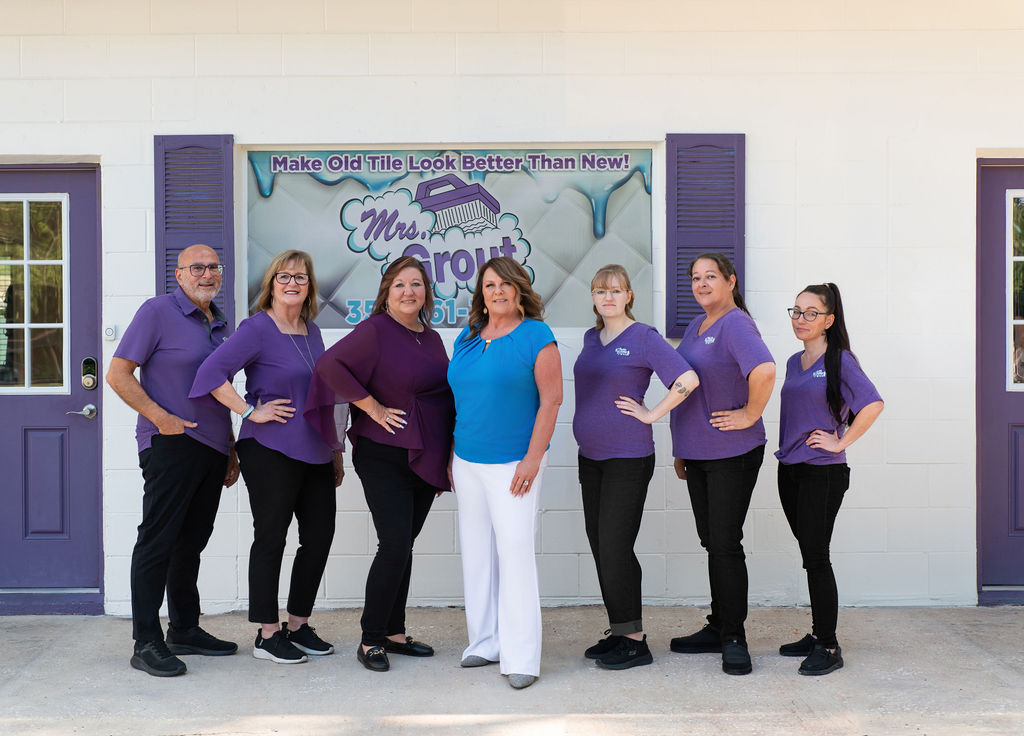 Mrs. Grout team members standing together outside their office in front of the company sign, smiling for a group photo. The image introduces the professional team behind Mrs. Grout's tile and grout restoration services.