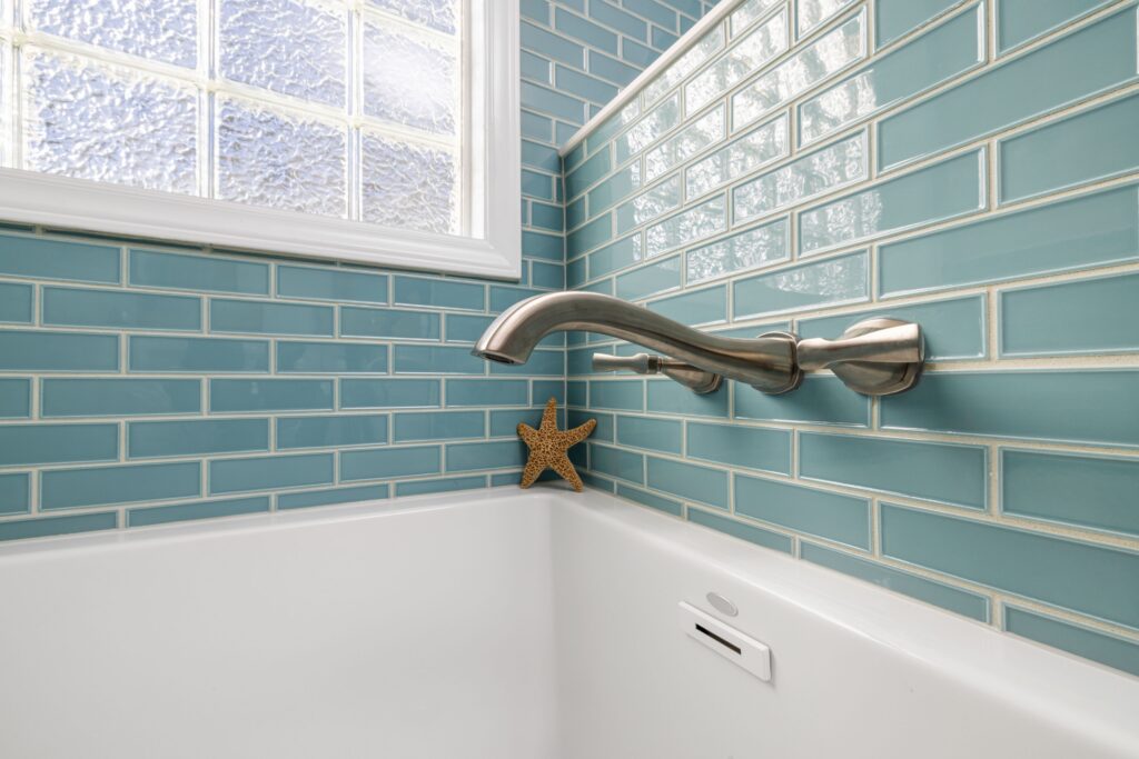 Clean bathroom with glossy blue subway tile walls, bright white grout lines, and a white bathtub beneath a frosted glass window. The image highlights a clean, well-maintained tiled bathroom.