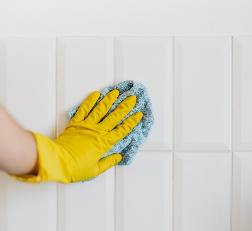 Close-up of a person wearing a yellow cleaning glove wiping white wall tiles with a blue microfiber cloth. The image shows routine tile cleaning and surface maintenance.