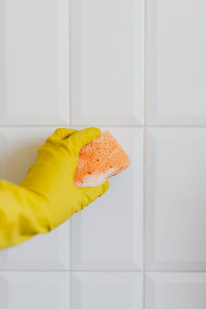 Hand wearing a yellow cleaning glove holding a sponge against clean white shower tiles. The image illustrates cleaning and maintaining tiled shower walls.