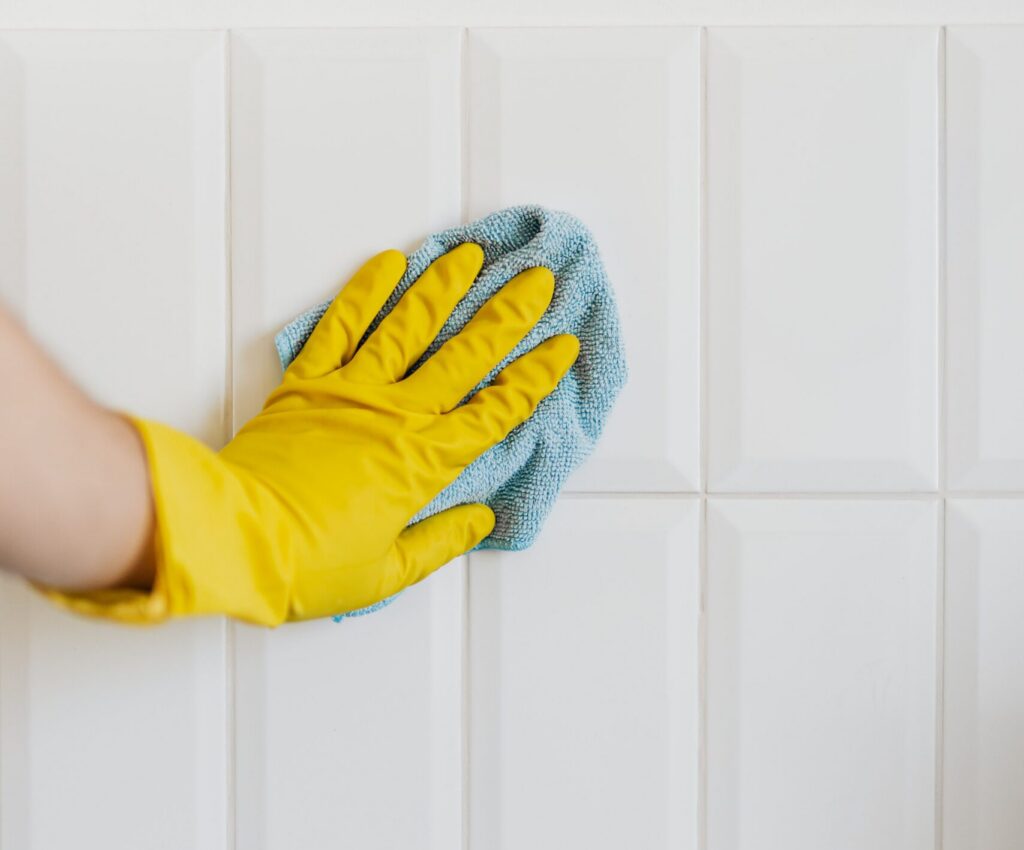 Close-up of a person wearing a yellow cleaning glove wiping white wall tiles with a blue microfiber cloth. The image shows routine tile cleaning and surface maintenance.