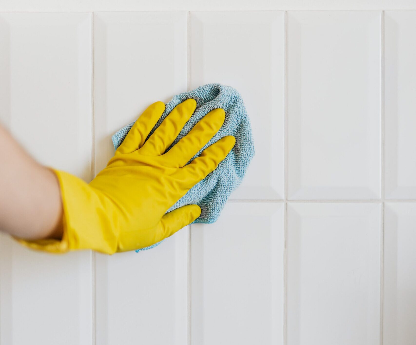 Close-up of a person wearing a yellow cleaning glove wiping white wall tiles with a blue microfiber cloth. The image shows routine tile cleaning and surface maintenance.