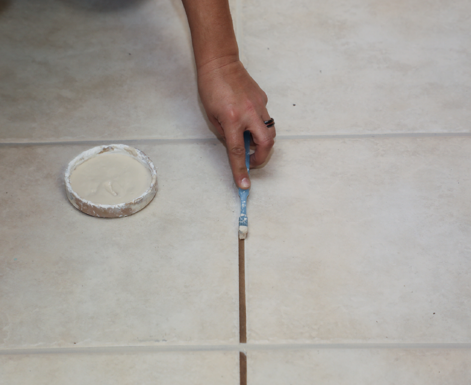 A person applies light-colored grout with a small brush to a vertical grout line between large beige ceramic floor tiles. An open container of grout sits beside the work area.