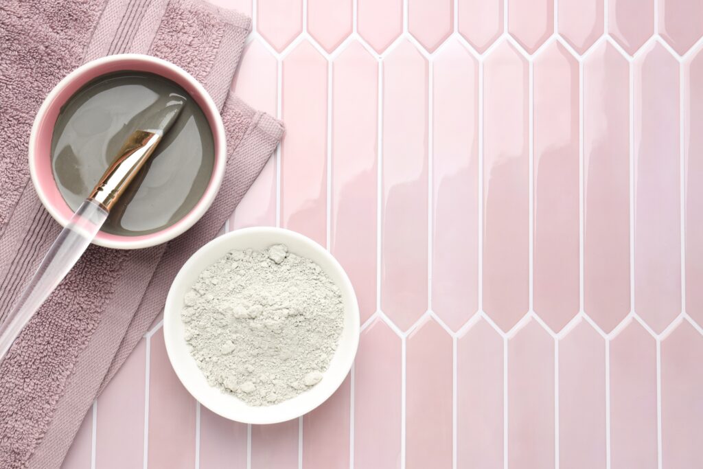 A cup with clay paste in it and a cup with mud and a brush in it, on top of a towel on pink tile.