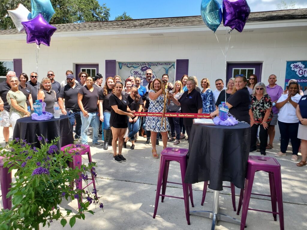 Group of employees and community members gathered outside a business for a ribbon-cutting ceremony celebrating a new Mrs. Grout location. Purple and blue balloons, ribbon-cutting supplies, and outdoor tables decorate the event.