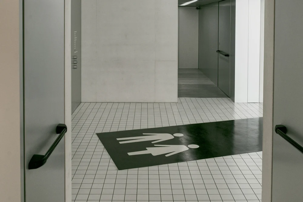 Modern hallway with open restroom doors leading to an all-gender restroom marked by a floor sign featuring universal person icons. The clean, minimalist space emphasizes accessibility, inclusion, and welcoming public facilities.