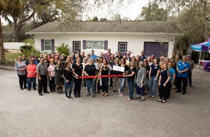 Group of employees and community members gathered outside a business for a ribbon-cutting ceremony celebrating a new Mrs. Grout location. Purple and blue balloons, ribbon-cutting supplies, and outdoor tables decorate the event.