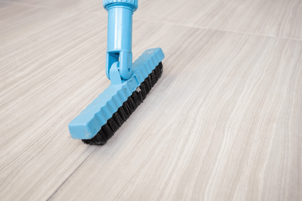 Close-up of a floor scrub brush cleaning a light-colored tile floor. The image illustrates routine floor cleaning and maintenance.