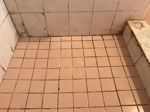 Close-up view of a tiled restroom floor and lower tiled wall showing visible wear, discoloration, and stained grout.