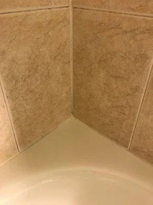 Close-up view of the corner of a tiled shower with light-colored wall tiles and a white shower base. The image shows grout lines and the condition of the shower surfaces.